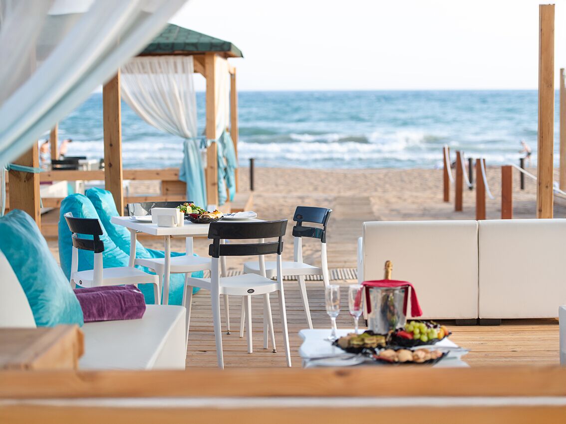 A beachside lounge with a wooden deck, white furniture, and a table set with food and drinks.