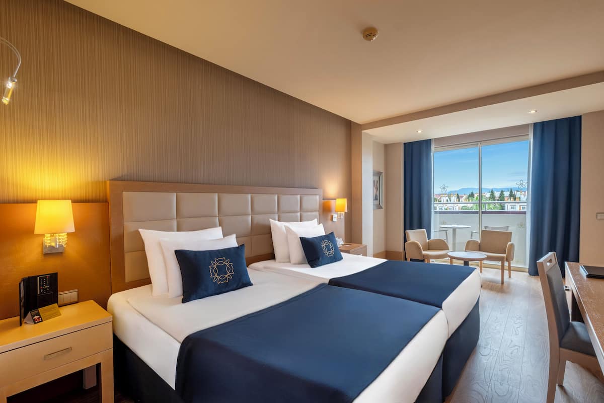 A hotel room with two beds, a desk, and a balcony with a view.
