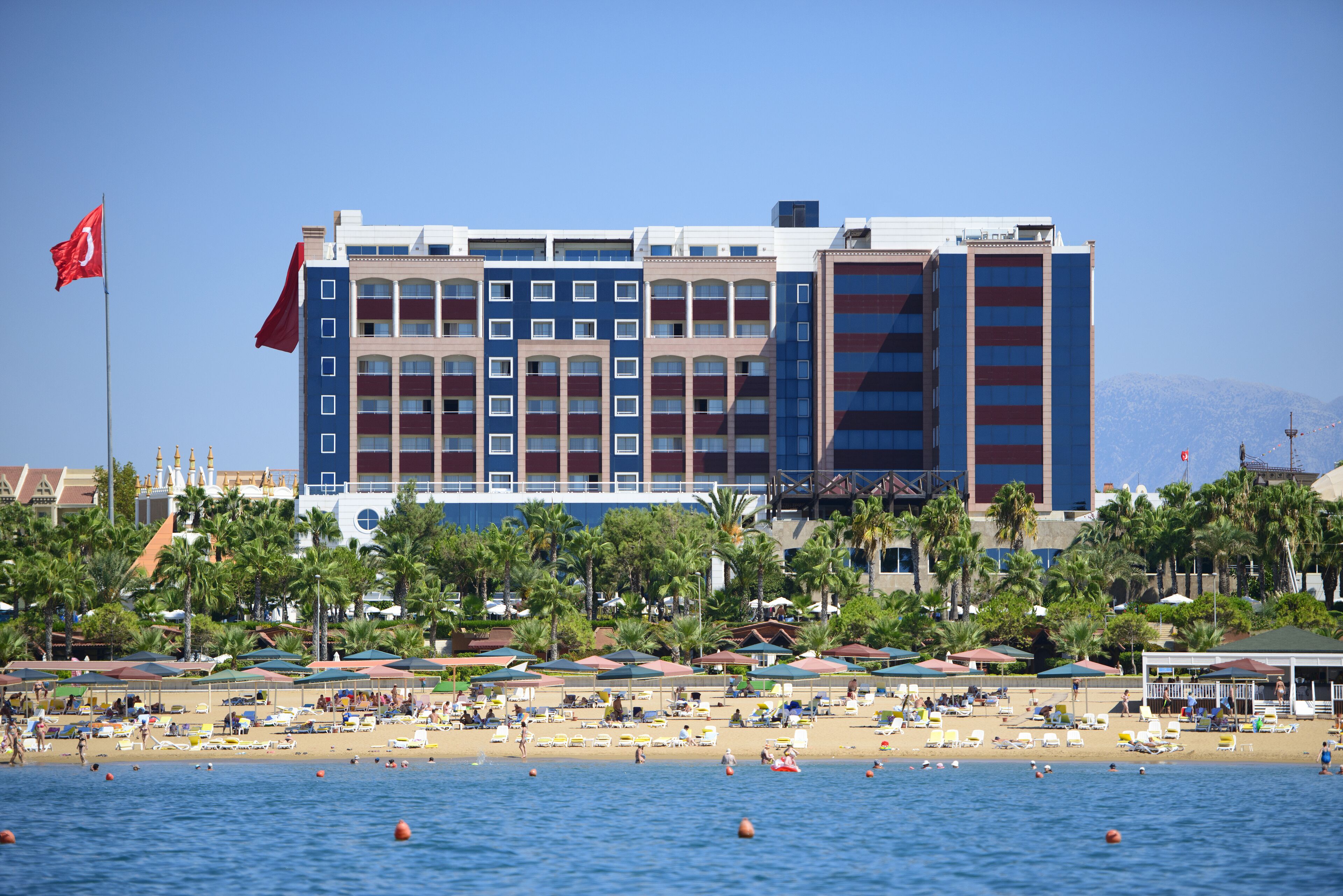 A hotel with multiple floors, a beach in the foreground, and palm trees.
