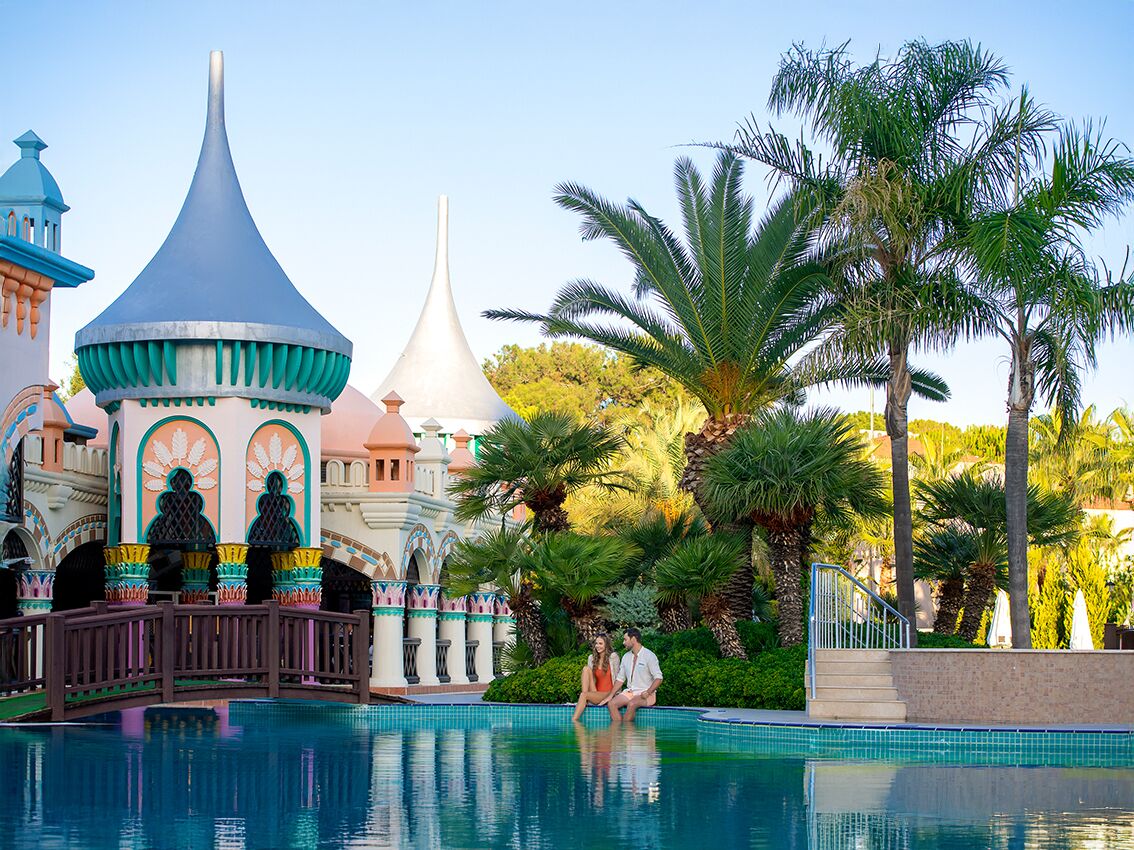 A pool area with a bridge, palm trees, and a building with a blue dome and white spire.