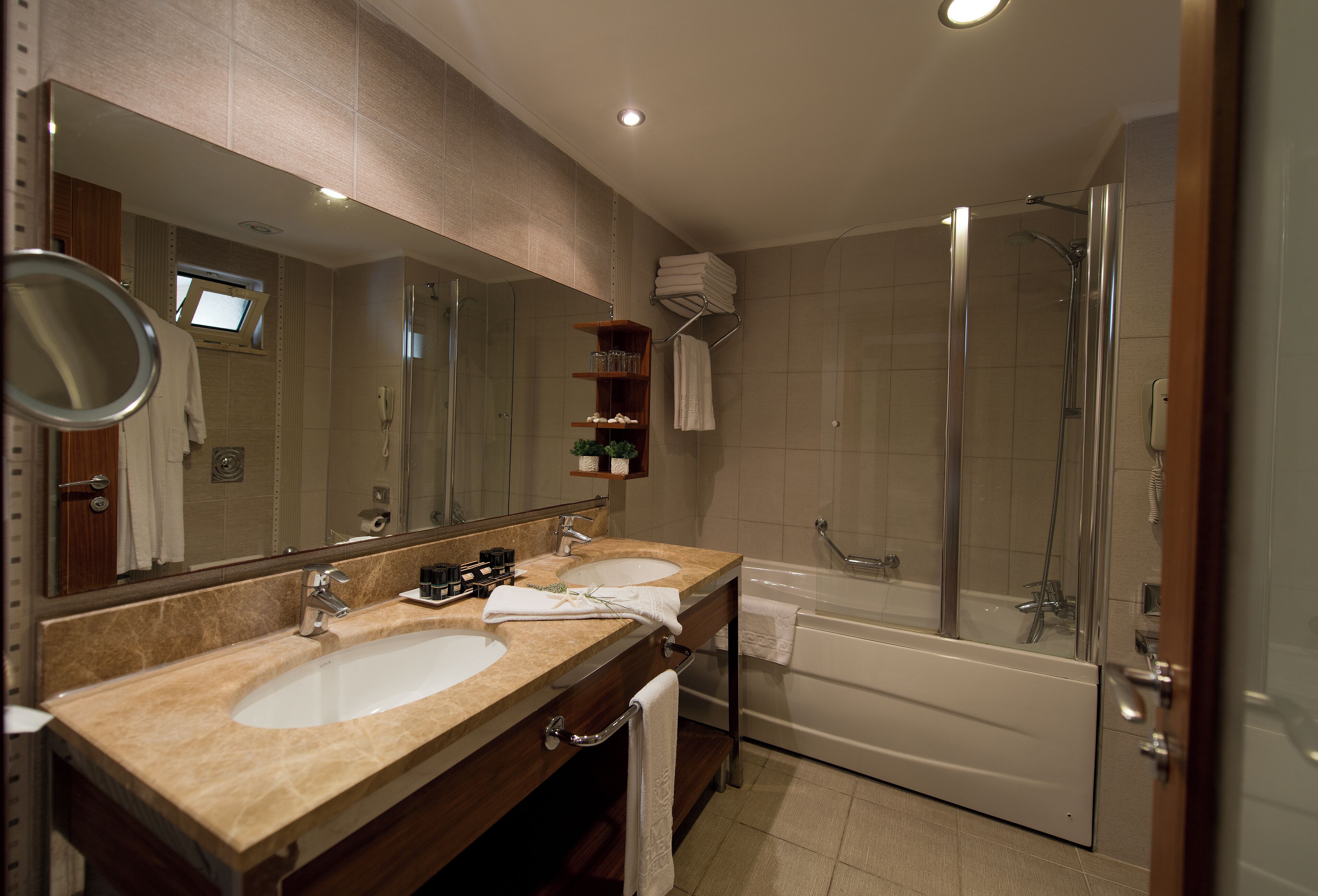 Un baño moderno con dos lavamanos, un espejo grande, ducha con mampara de vidrio y estante empotrado con artículos de aseo.