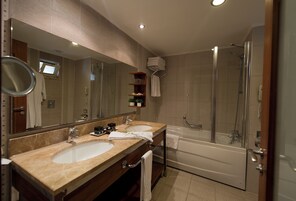 Un baño moderno con dos lavamanos, un espejo grande, ducha con mampara de vidrio y estante empotrado con artículos de aseo.