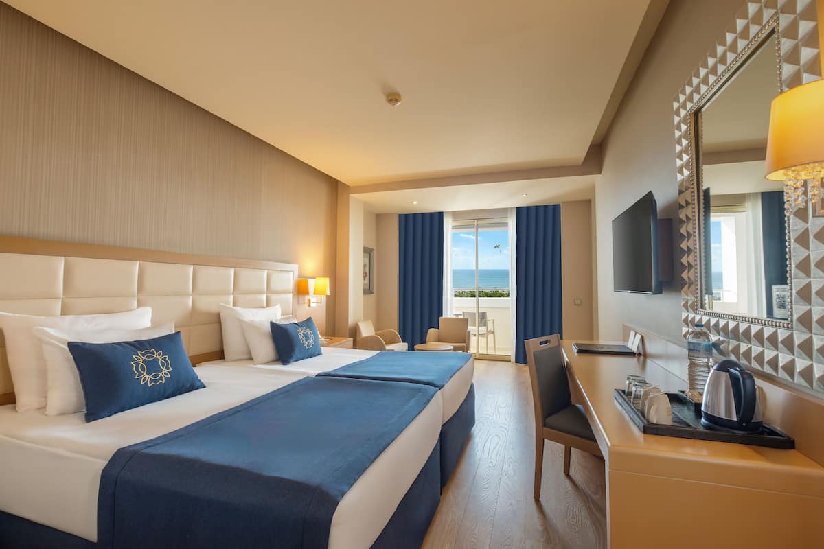 A hotel room with two beds, a desk, a chair, a TV, and a view of the sea.