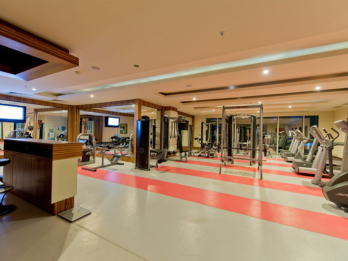 Une salle de sport équipée de divers appareils d’exercice, un comptoir d’accueil et un sol aux motifs rouges et blancs.