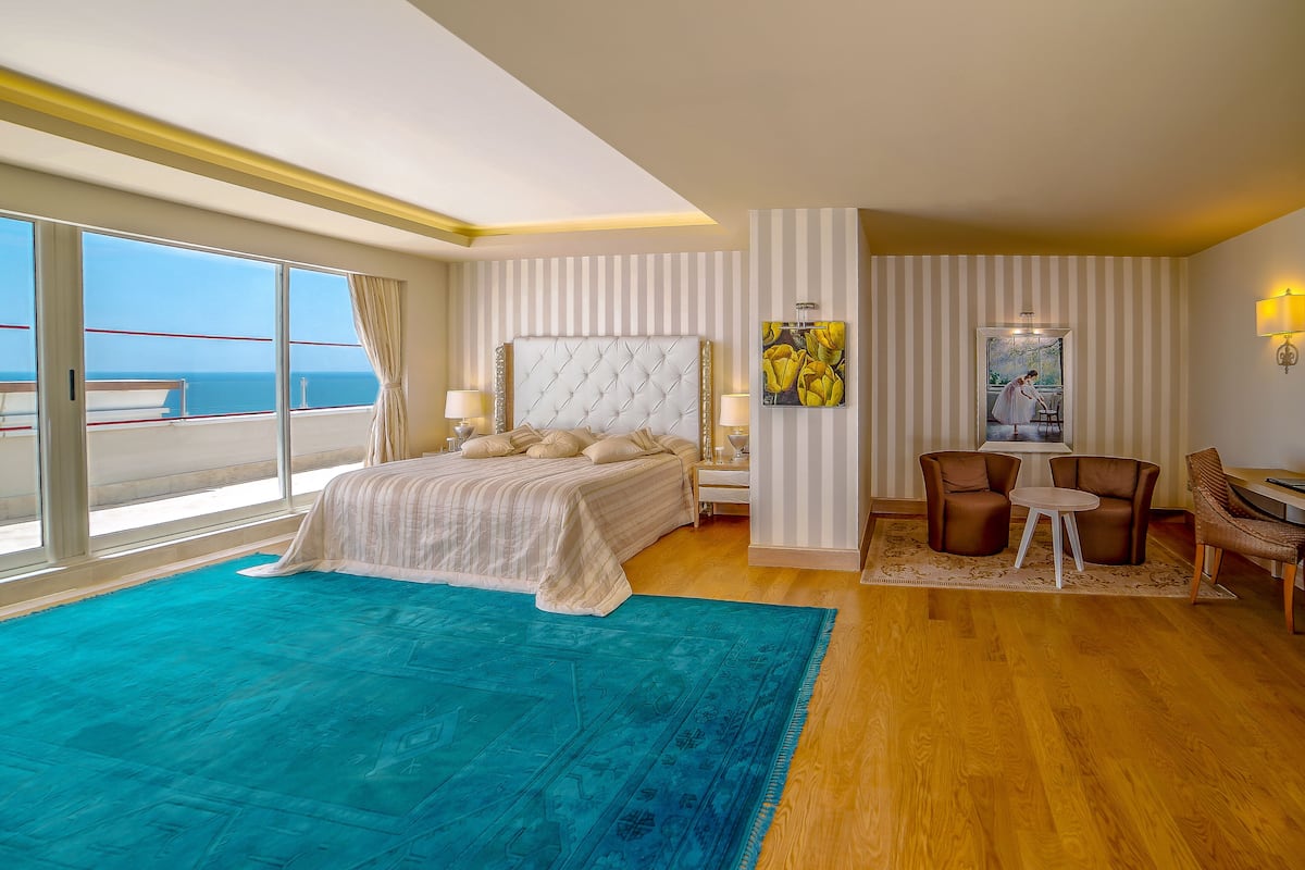A spacious bedroom with a large bed, a sitting area with chairs, a view of the sea through large windows, and a balcony.