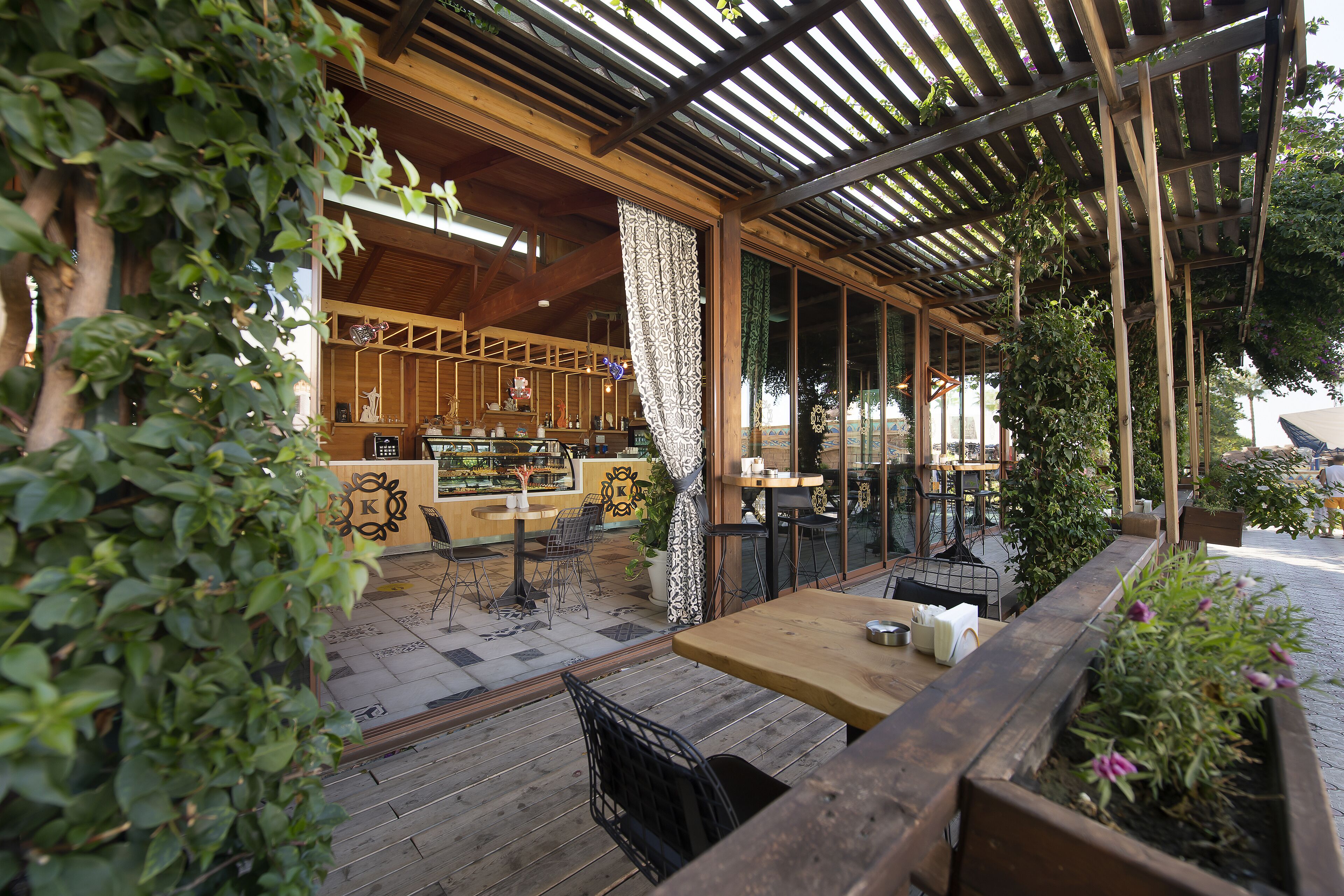 Een openlucht café met houten tafels en stoelen, een toonbank met een decoratief logo, en een pergola met klimplanten.