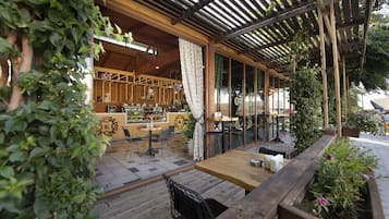 Een openlucht café met houten tafels en stoelen, een toonbank met een decoratief logo, en een pergola met klimplanten.