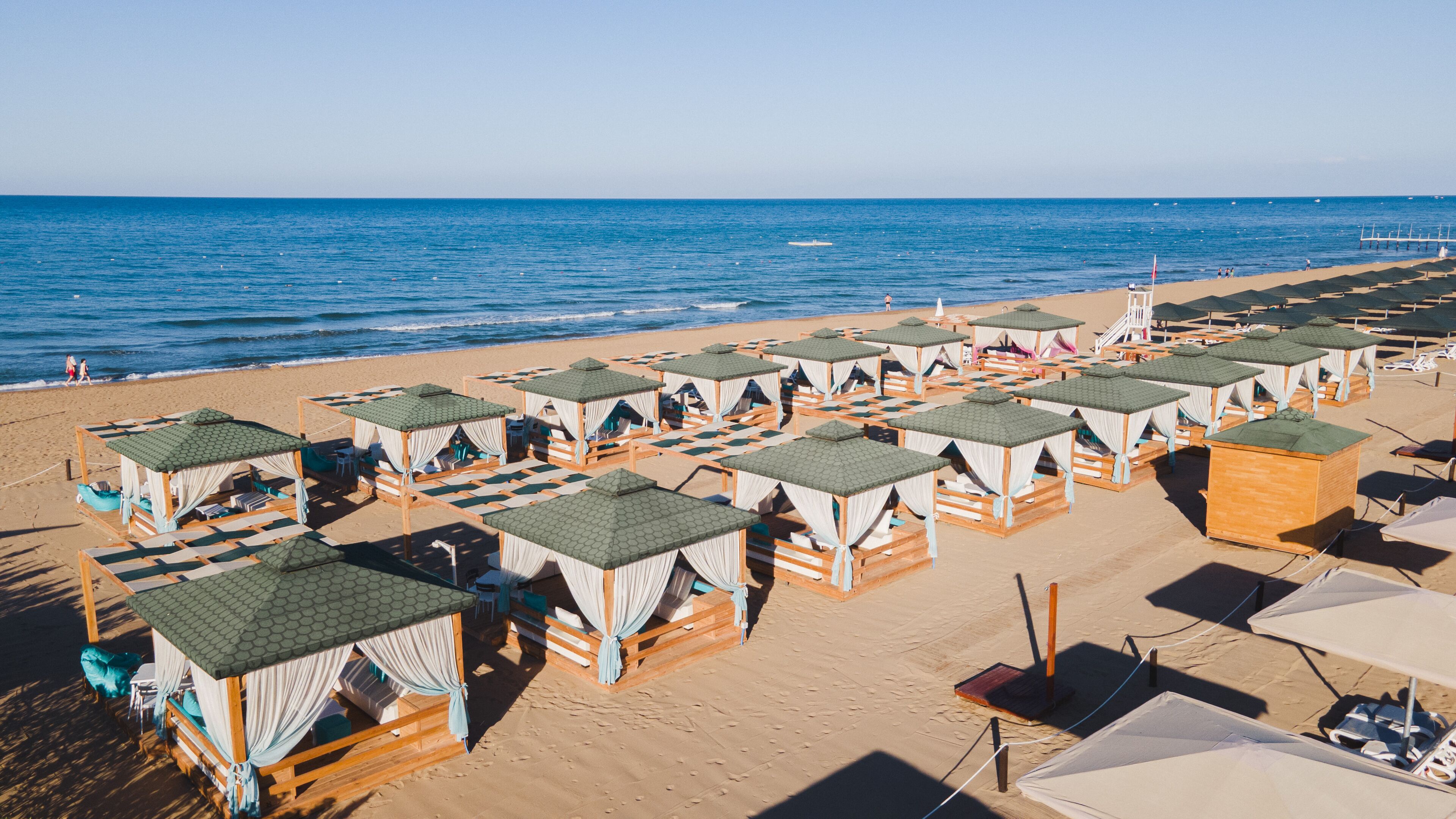 A beach with rows of wooden cabanas with green roofs and white curtains, arranged on the sand.