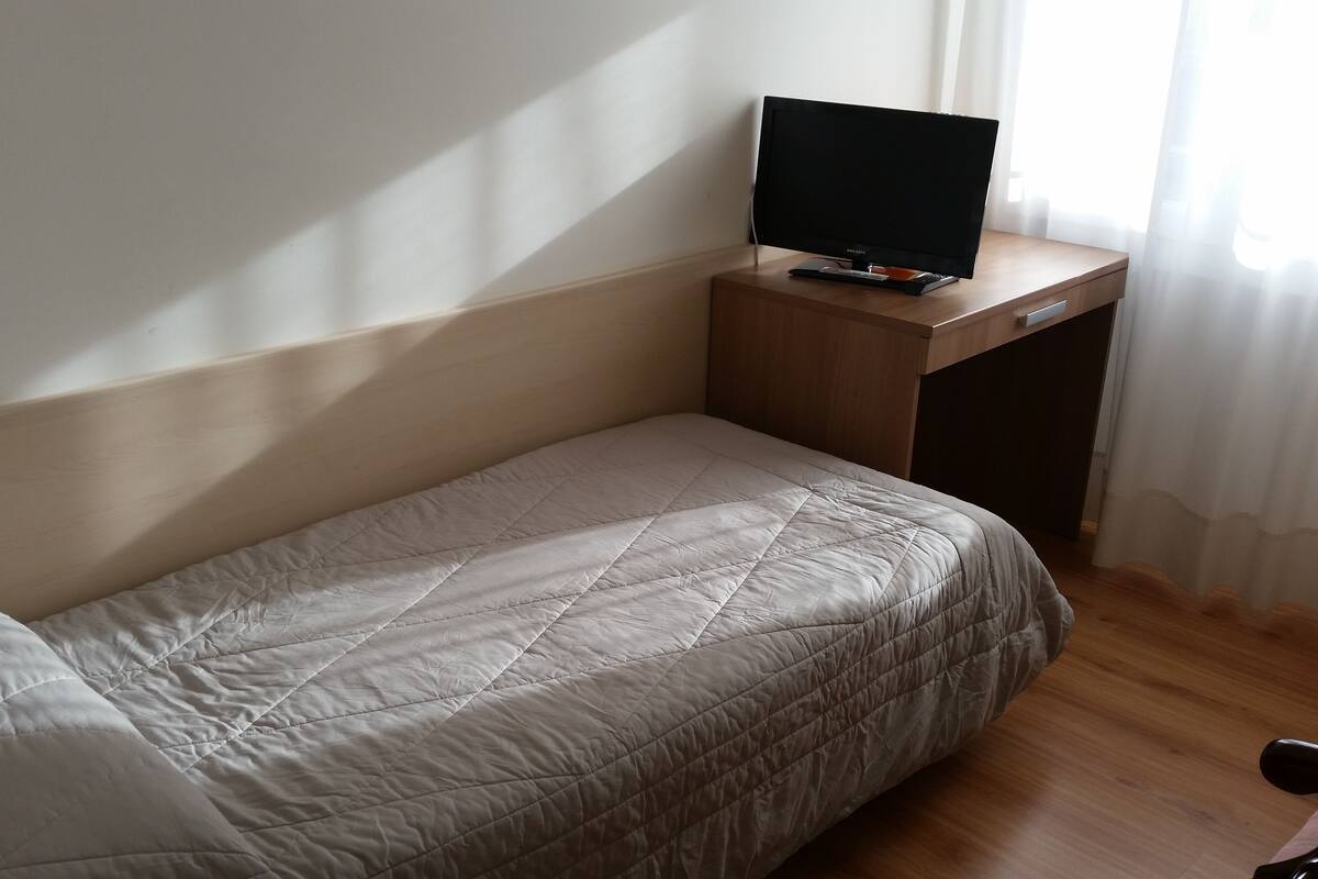 Ein Einzelbett mit einer Steppdecke, ein Holztisch mit Fernseher und ein Fenster mit Vorhängen.
