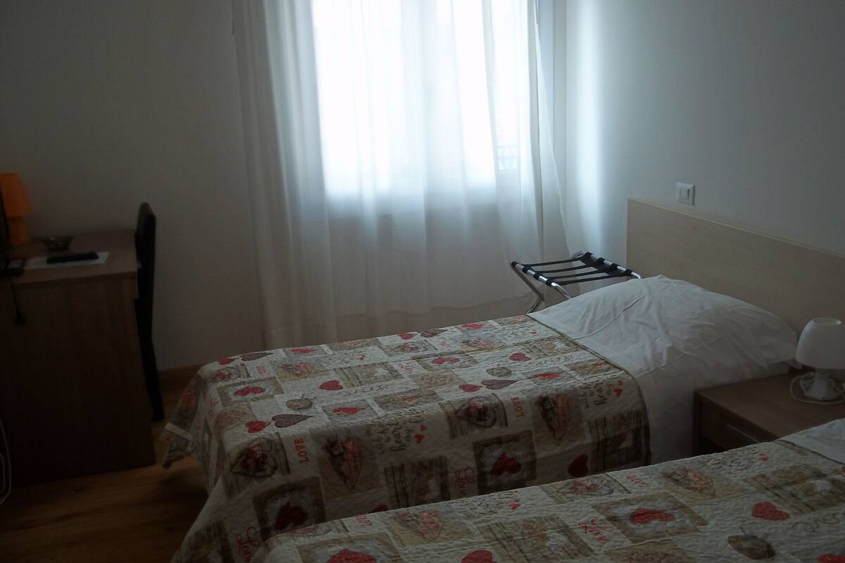 Ein Schlafzimmer mit zwei Betten, einem Nachttisch, einer Lampe und einem Fenster mit durchsichtigen Vorhängen.