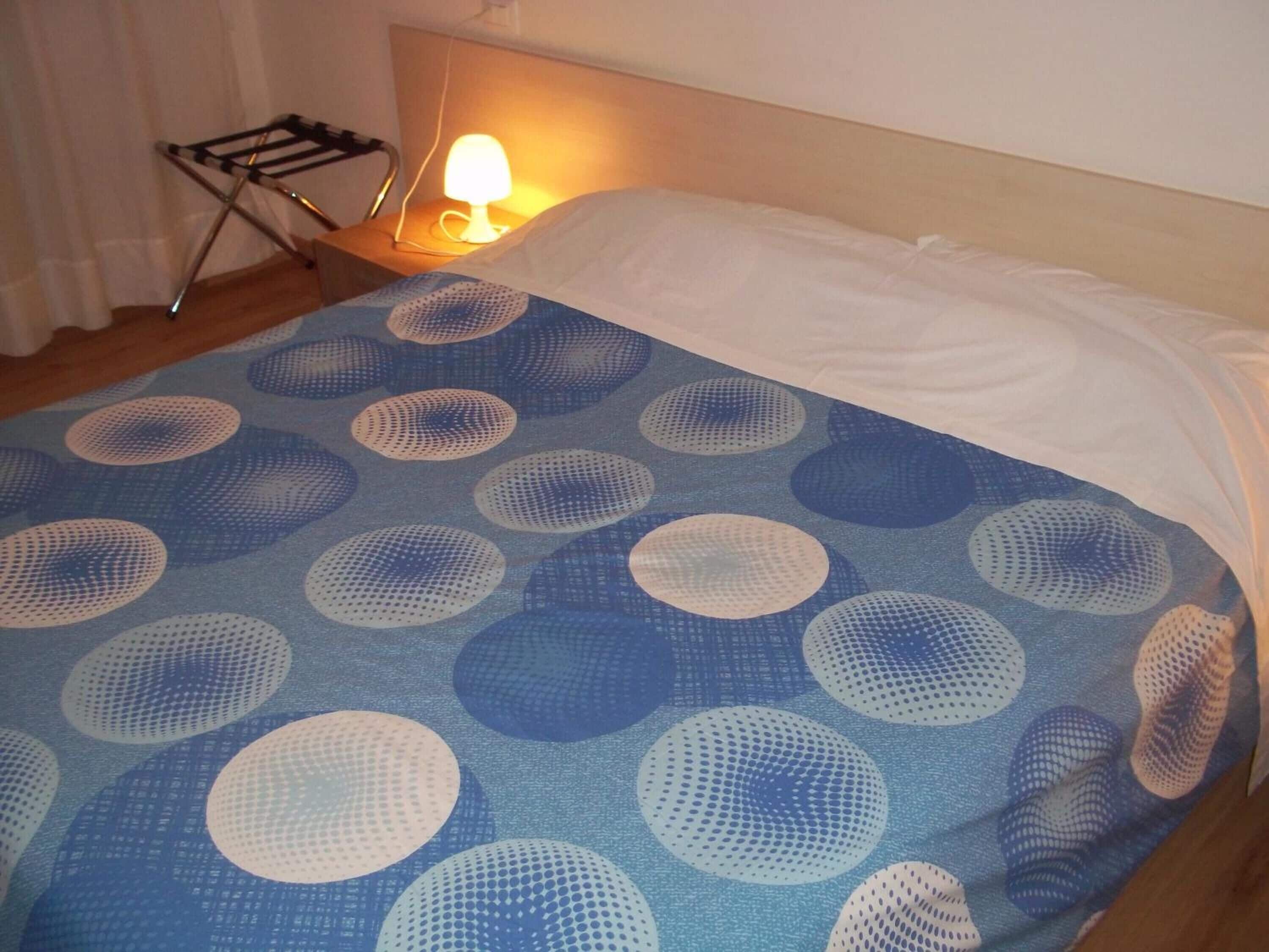 Un lit recouvert d’une couvre-lit aux motifs circulaires bleus et blancs, un oreiller blanc et une lampe de chevet posée sur une table de nuit en bois.
