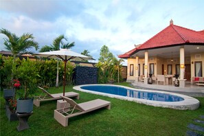 A swimming pool with lounge chairs and umbrellas, surrounded by a well-maintained lawn and tropical plants. - Bali Asih Villa (Kerobokan)