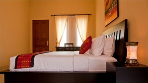 Een net opgemaakt bed met witte lakens en een rode decoratieve kussen, een houten nachtkastje met een lamp, en een deur met gordijnen in een kamer met beige wanden.