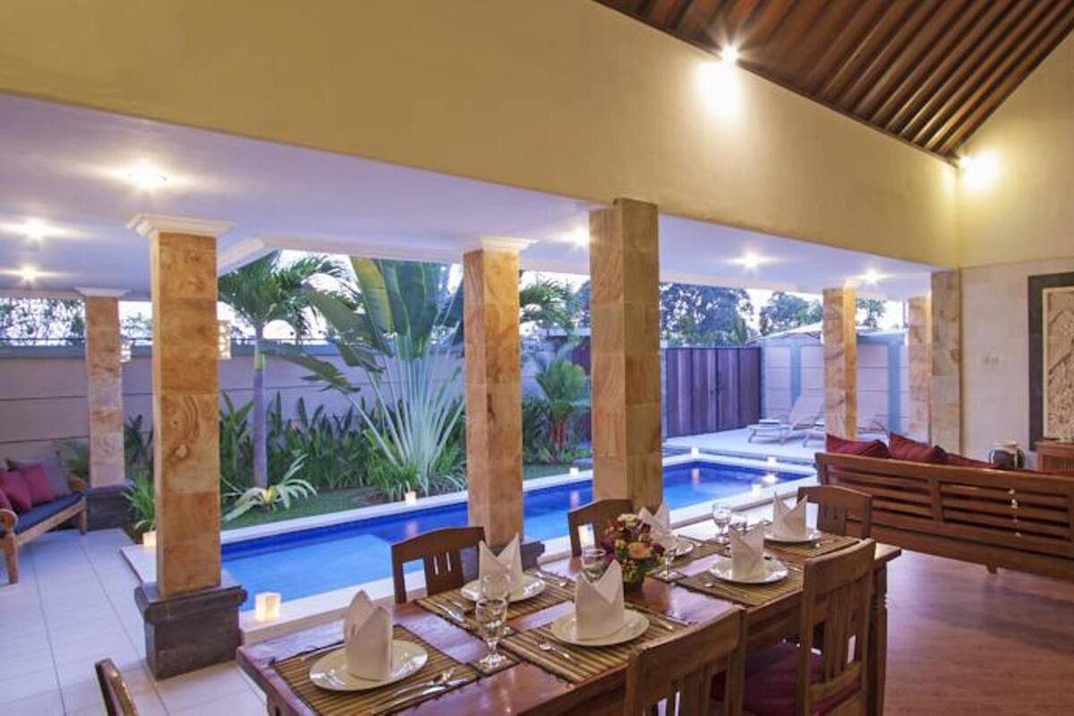 A dining area with a pool view, wooden furniture, and a TV in the background.