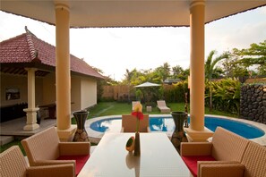 A covered outdoor area with a pool, wicker furniture, and a table with a vase. - Bali Asih Villa (Kerobokan)