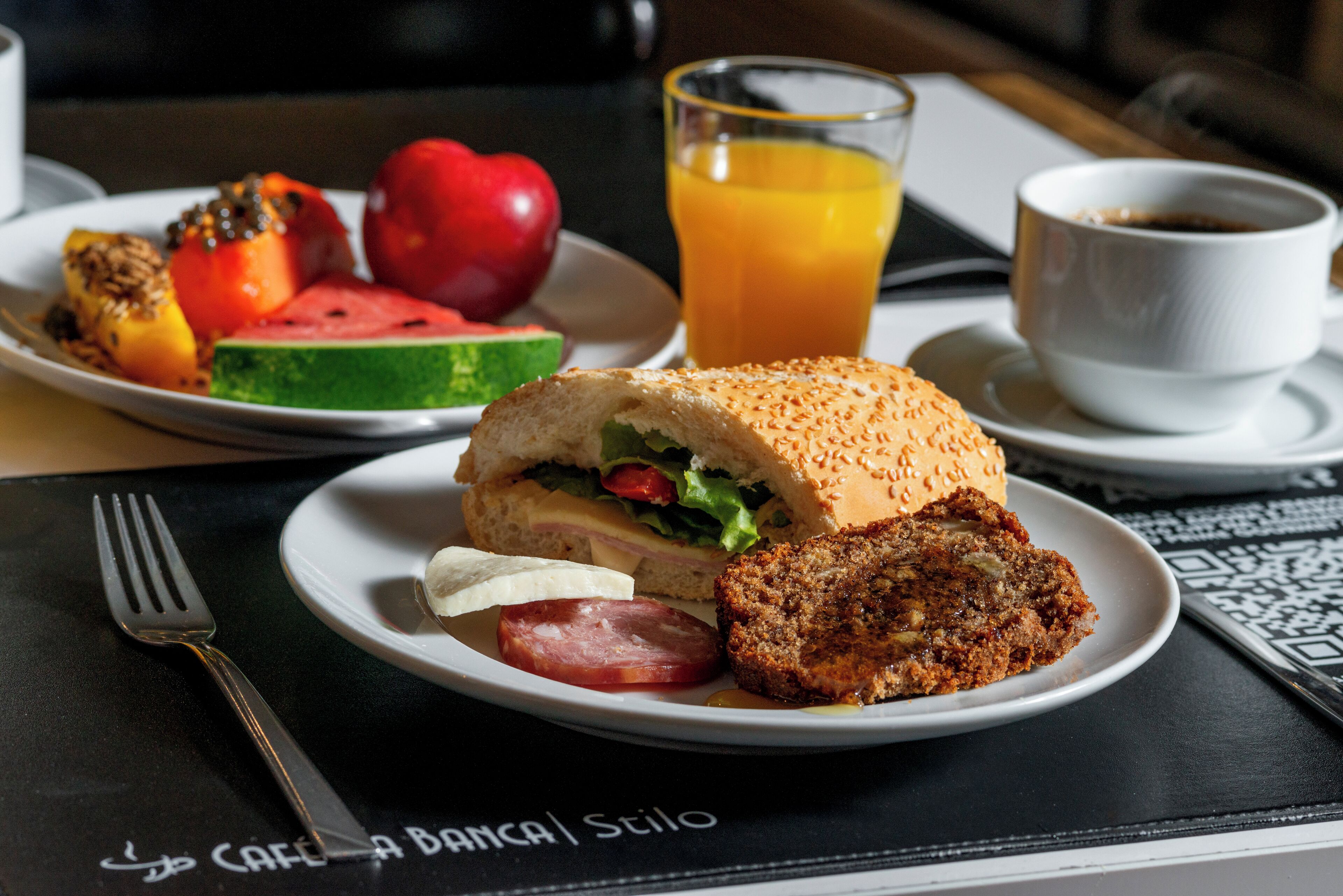 Un plato con un sándwich, una porción de carne, una rodaja de tomate, una tajada de sandía y un vaso de jugo de naranja.