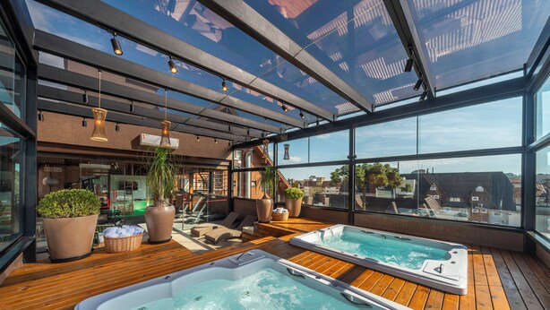 A rooftop terrace with two hot tubs, wooden flooring, potted plants, and a glass ceiling offering a city view. - Hotel Laghetto Stilo Centro (Gramado)