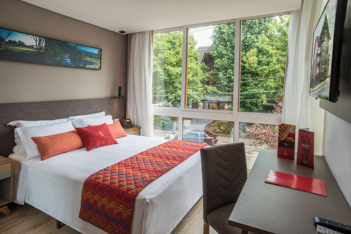 Ein Hotelzimmer mit einem großen Bett, einem Schreibtisch und Blick auf Bäume aus dem Fenster.