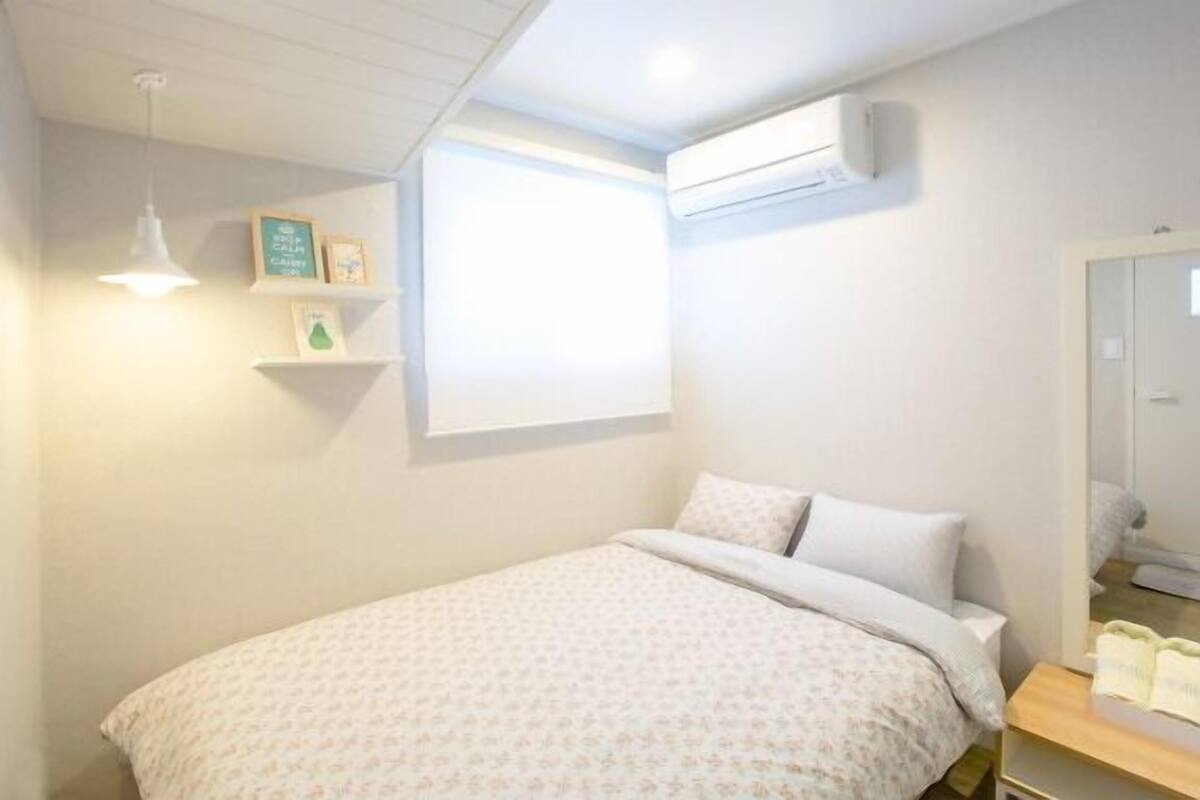 A bedroom with a bed, a bedside table, a wall-mounted air conditioner, and a window with blinds.