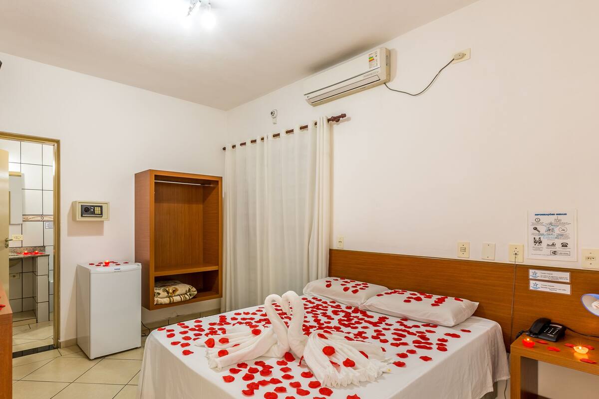 Una camera d'albergo con un letto ricoperto di petali di rosa, un minibar, un armadio in legno, un bagno con doccia e una televisione a parete.