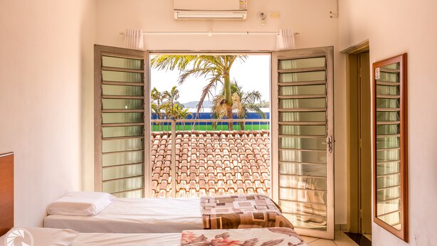 A hotel room with a bed, a small table, and a view of palm trees and a blue sky through open shutters. - Pousada das Estrelas Atibaia (Atibaia)