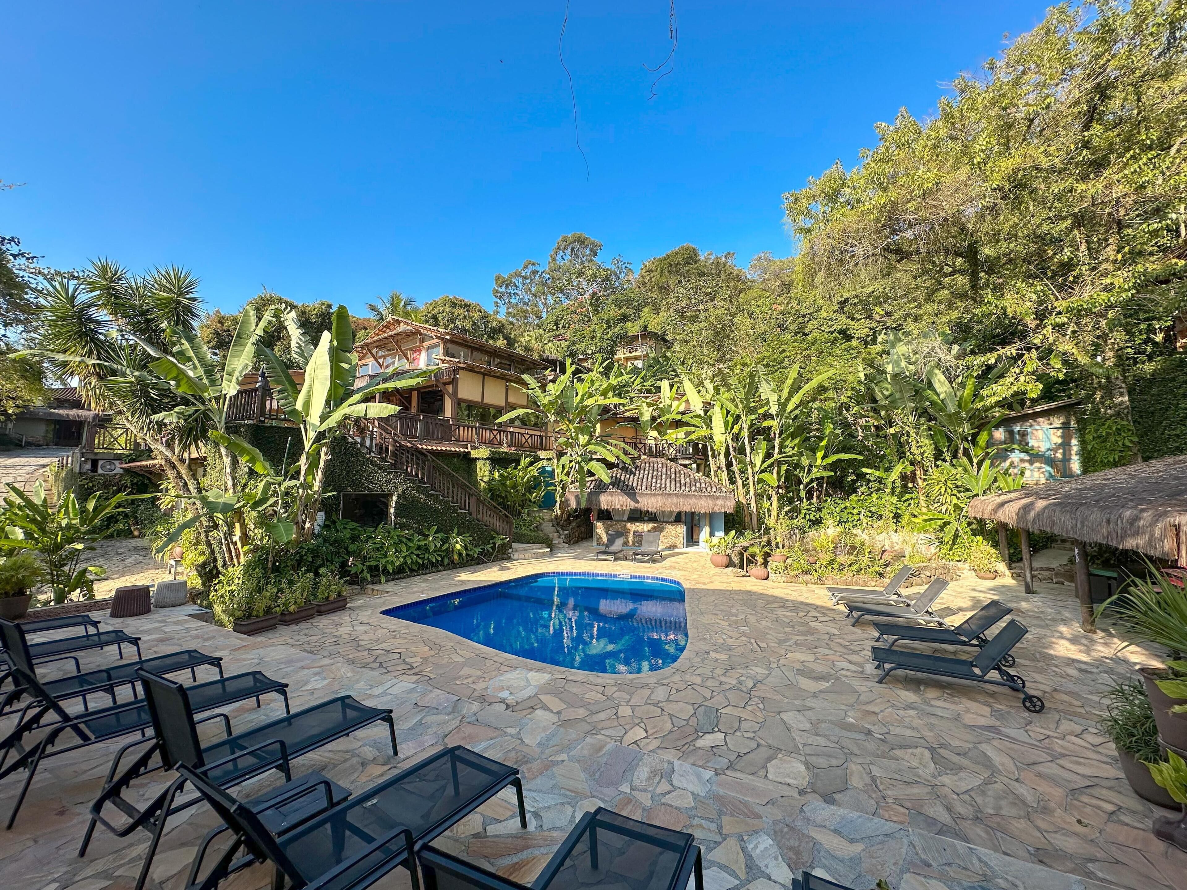 Un área de piscina con suelo de piedra, sillones de descanso y una casa al fondo.