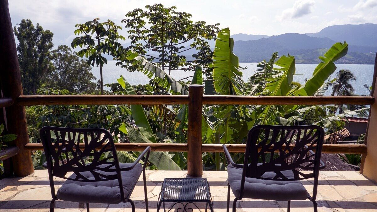 A balcony with two chairs and a small table, overlooking a scenic view of mountains and a body of water, surrounded by lush greenery.