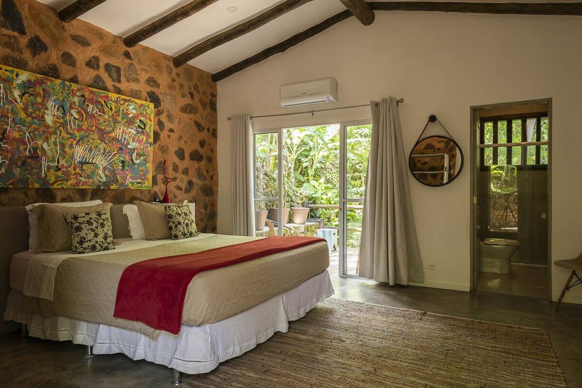 Een slaapkamer met een groot bed, een stenen muur, houten balken, een groot raam en uitzicht op groen.