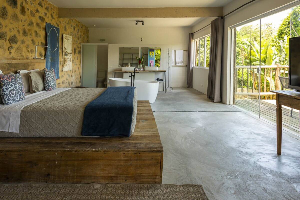 Een moderne slaapkamer met een groot bed, een houten bankje en uitzicht op een badkamer en balkon.