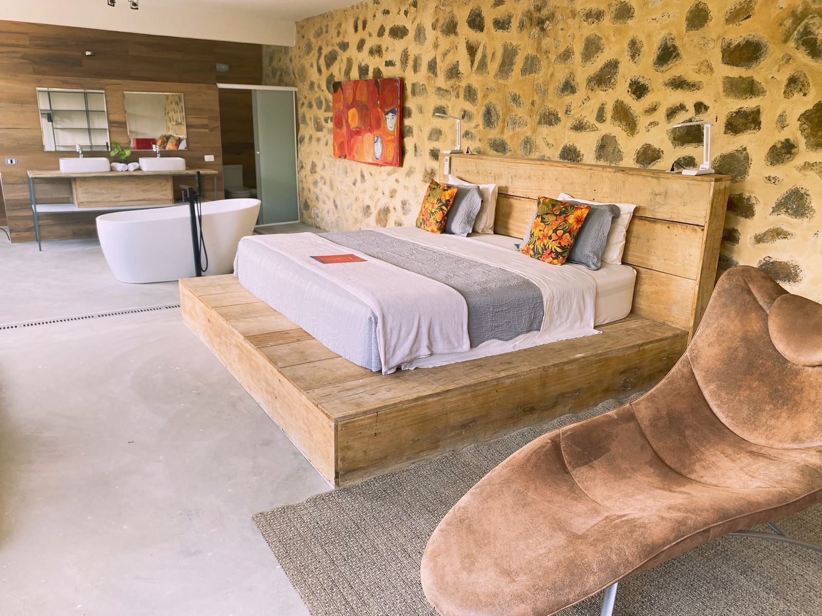 Un dormitorio moderno con una cama de madera, un espejo grande, una pared de piedra y un baño con lavamanos y bañera blancos.