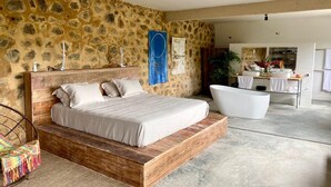 Un dormitorio con pared de piedra, cama de madera, bañera blanca y baño con lavabo y espejo.