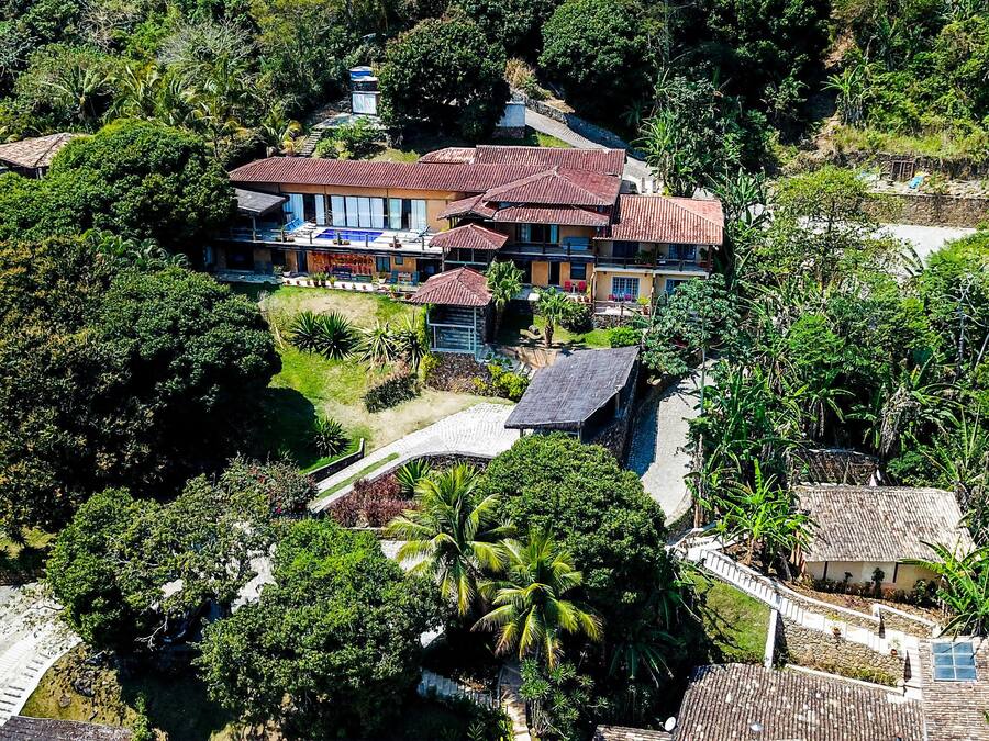 Vista aérea de uma casa grande, com vários andares e telhado de telhas vermelhas, cercada por vegetação exuberante e um caminho de pedra que leva à entrada.