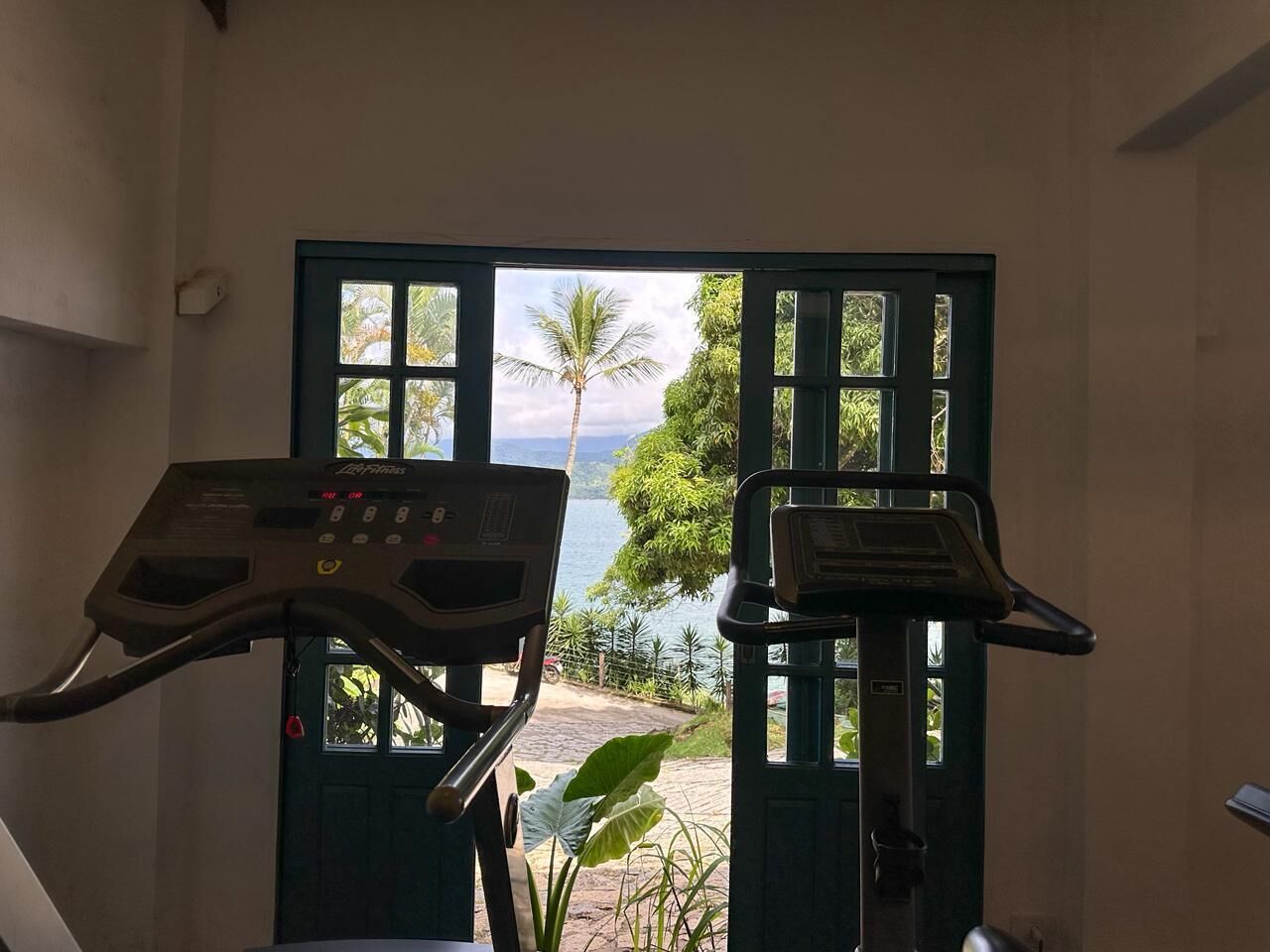 An indoor exercise area with two stationary bikes, a door leading to a scenic outdoor view with palm trees and a body of water.