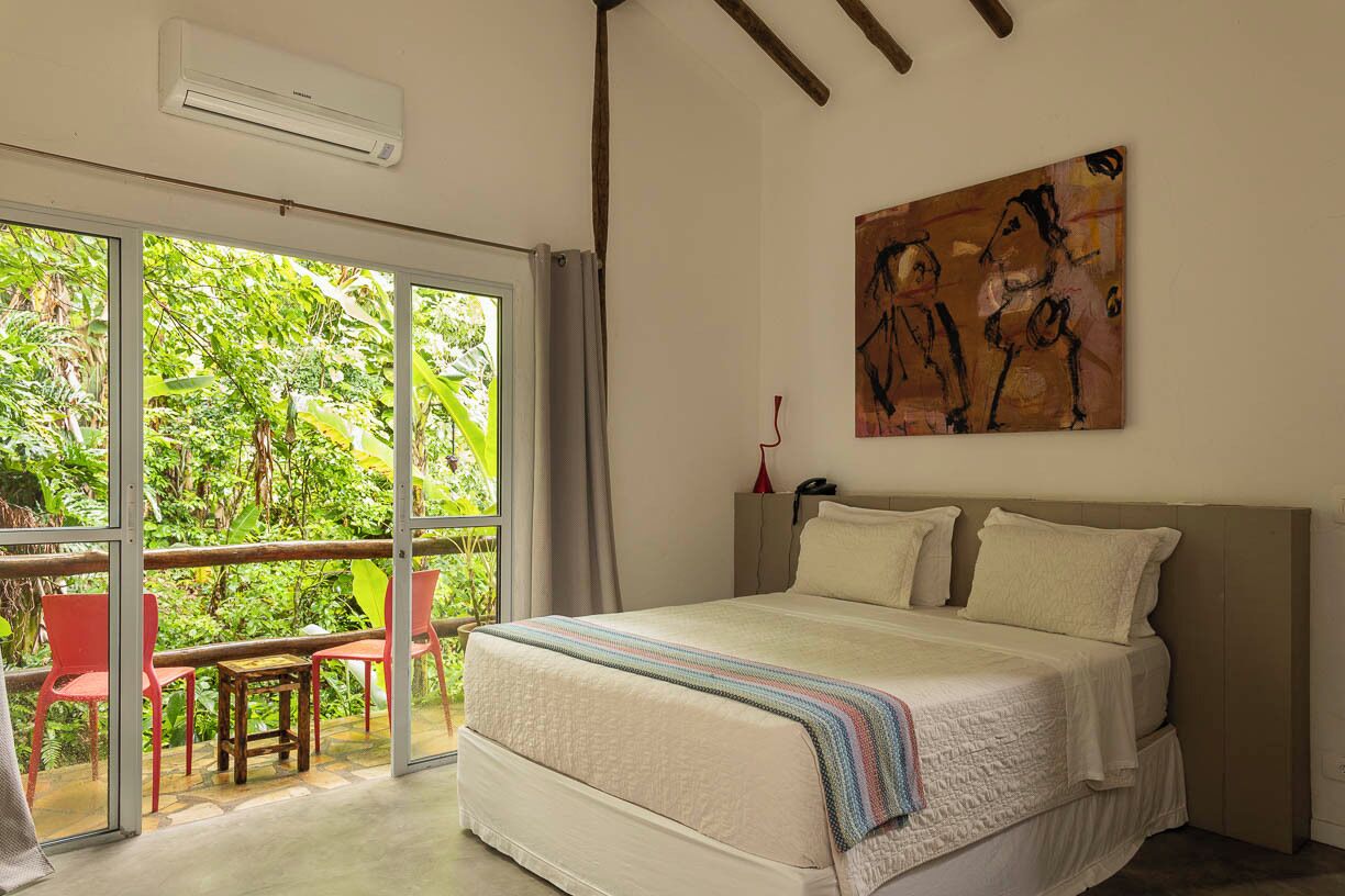 Un dormitorio con una cama grande, un ventanal corredero que da a un espacio exterior con sillas y un cuadro en la pared.
