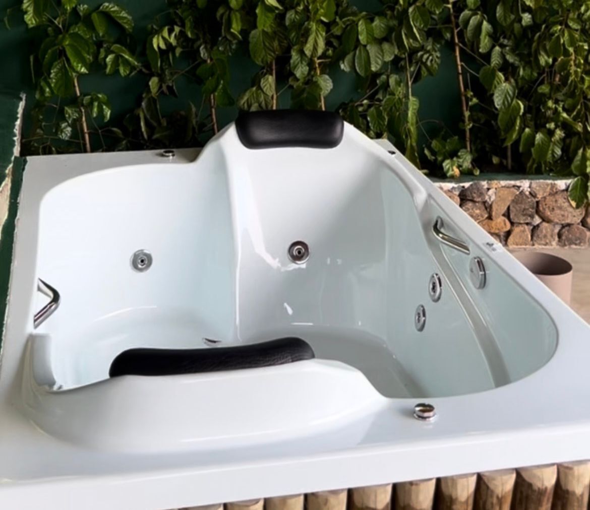 A white hot tub with a black seat, set against a backdrop of green foliage and a stone wall.