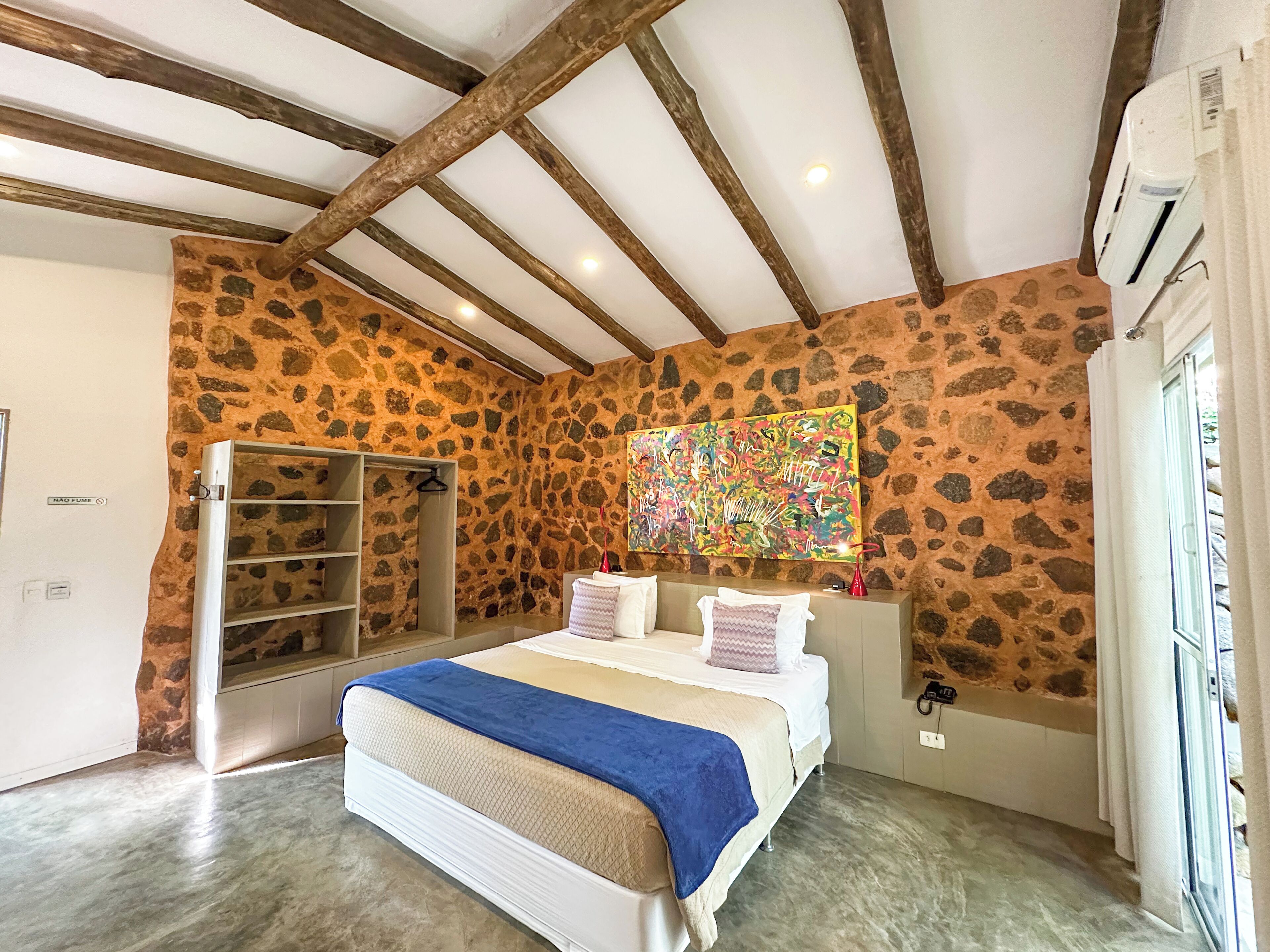 Un dormitorio con pared de piedra, cama con manta azul, techo de madera y un cuadro abstracto de colores.