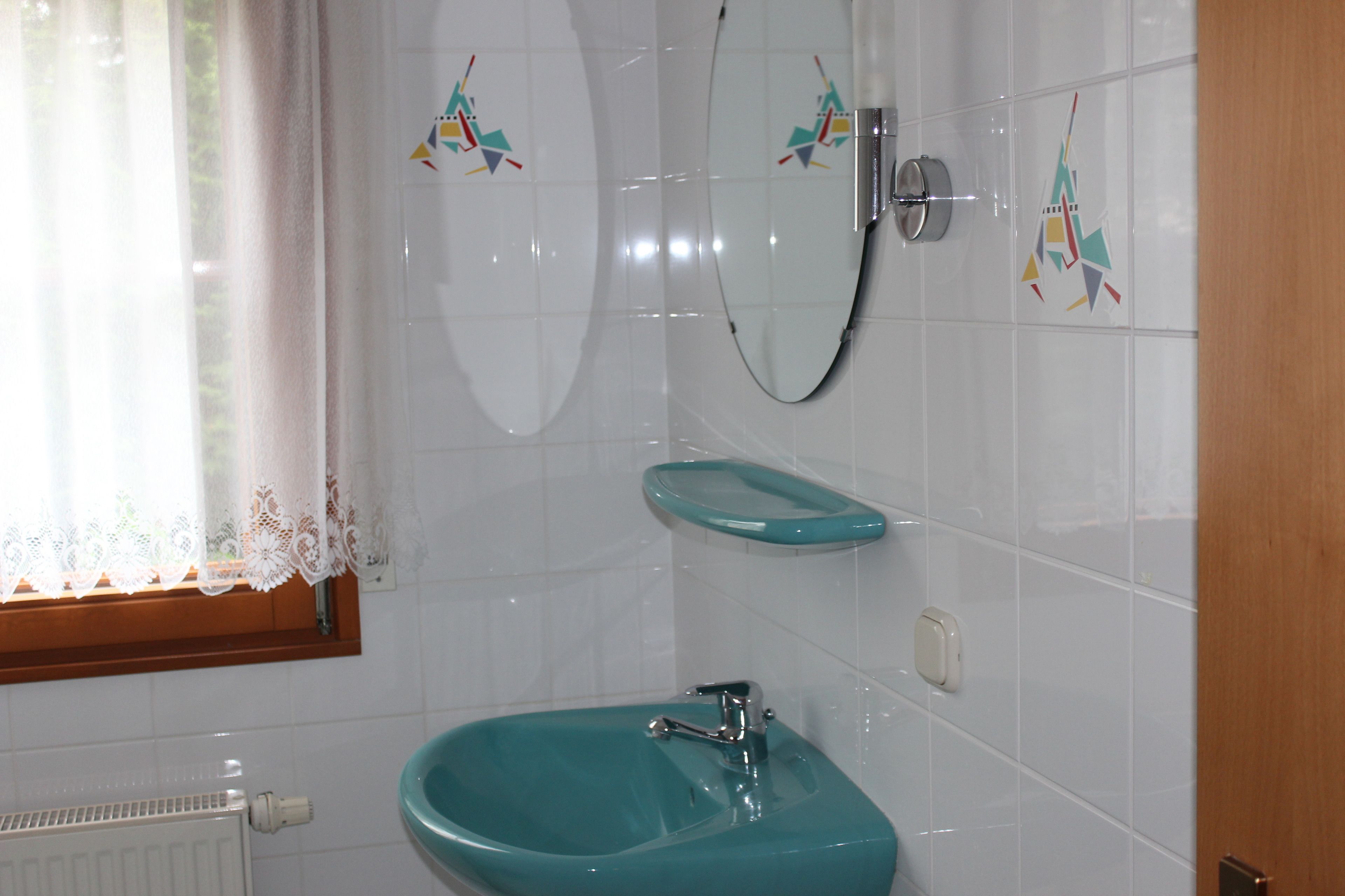 Une salle de bain avec un lavabo turquoise, un miroir et une fenêtre ornée de rideaux en dentelle.