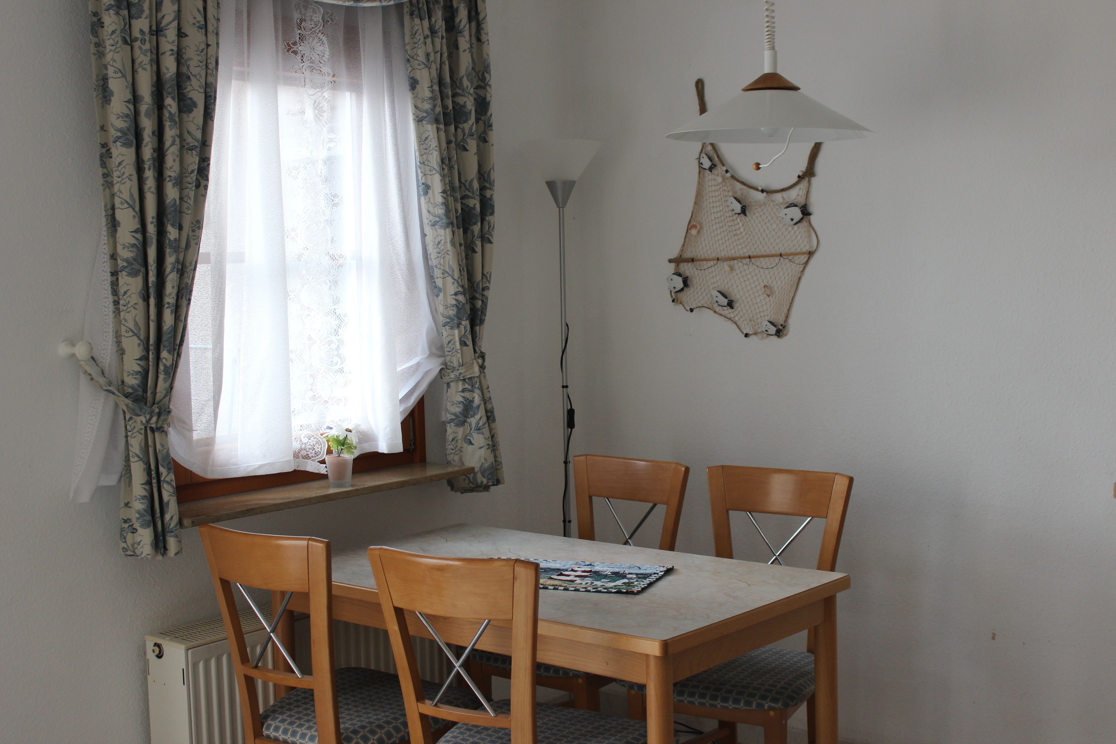 Zona pranzo con tavolo, quattro sedie di legno, lampadario e finestra con tende a fiori.