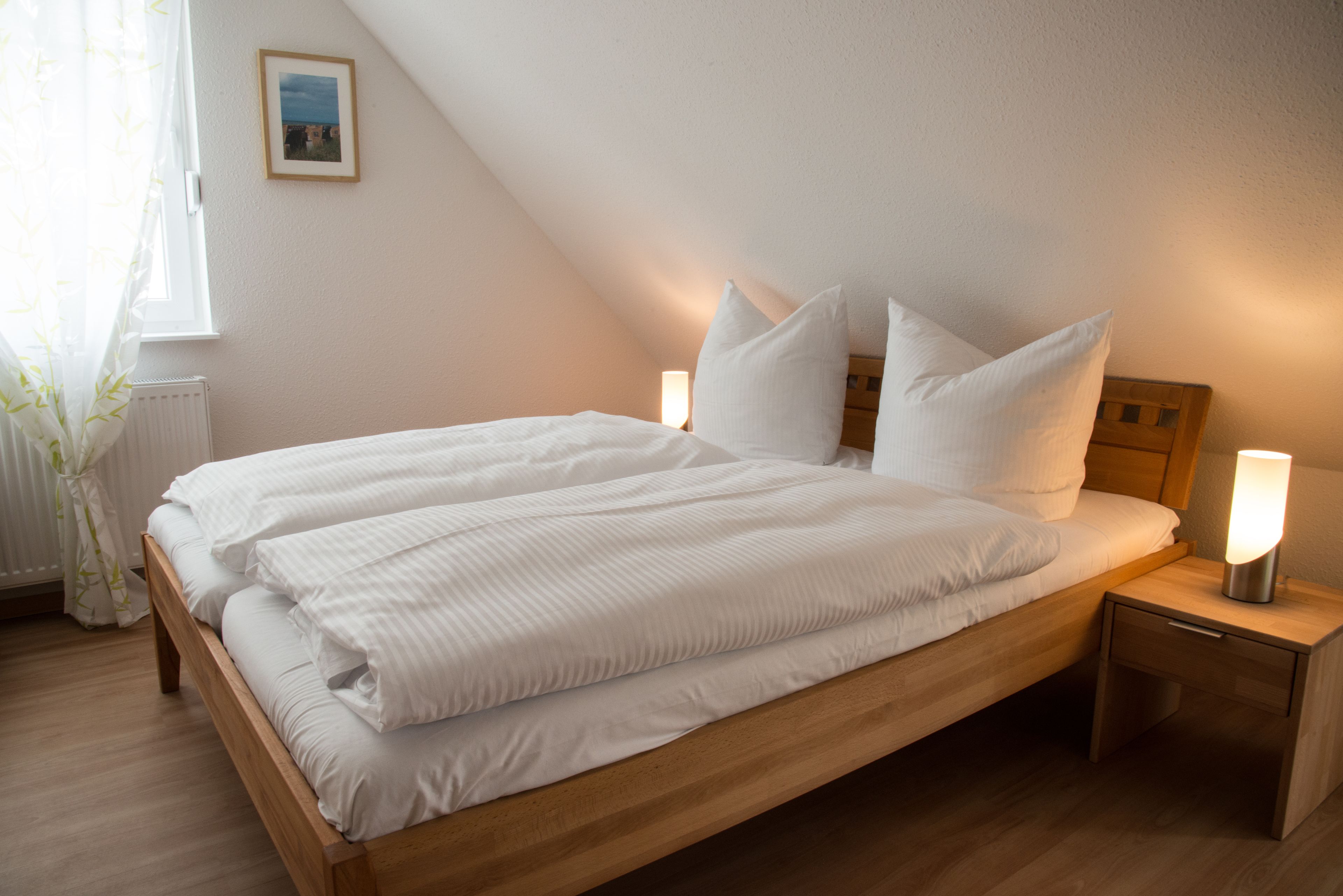 Una cama bien tendida con sábanas y almohadas blancas, una mesita de noche con una lámpara y un cuadro en la pared.