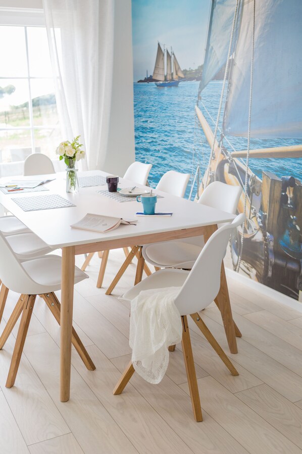 A dining area with a white table, chairs, and a view of a sailboat on the water through a large window.