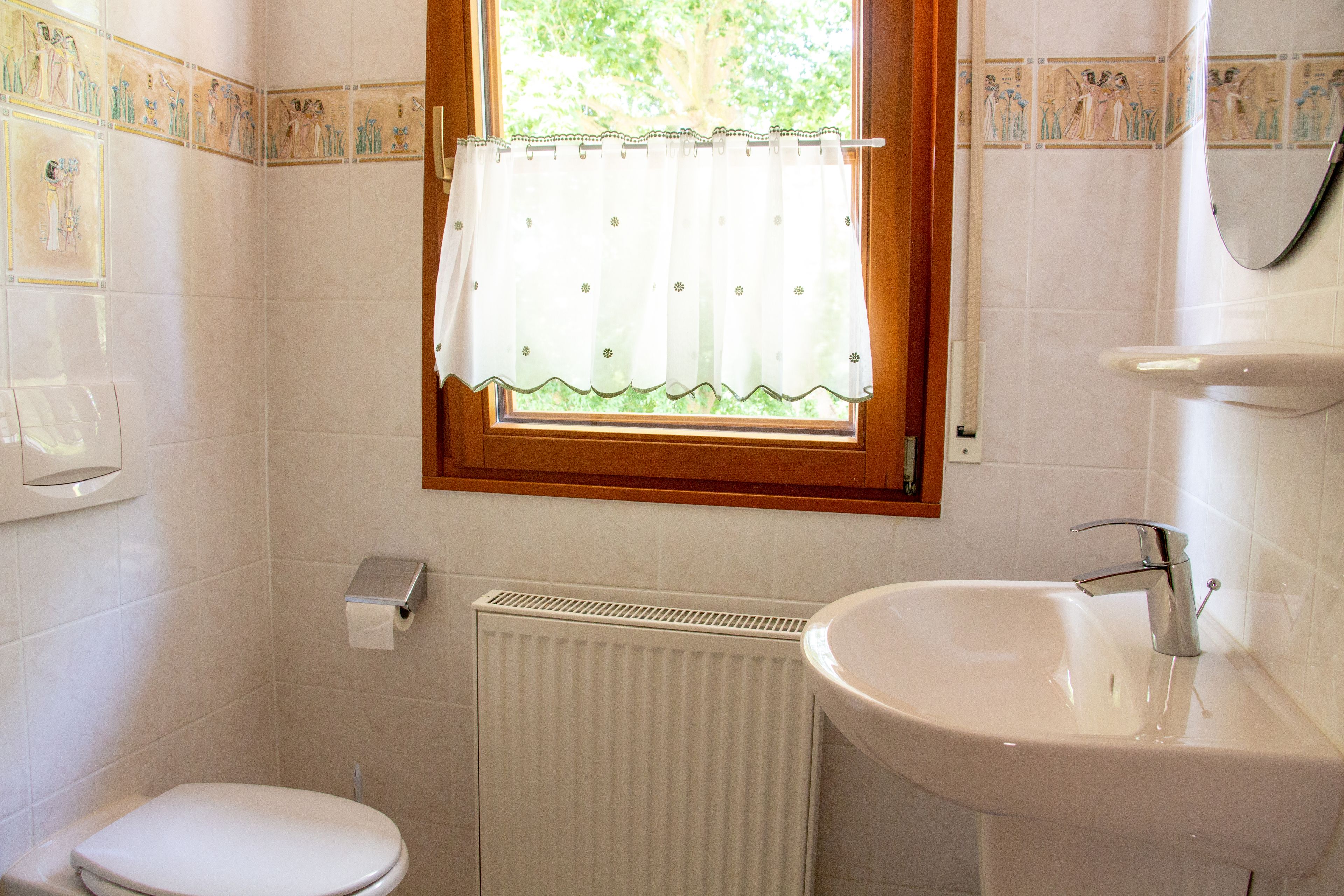 Um banheiro com vaso sanitário, pia, janela com cortina e azulejos decorativos na parede.