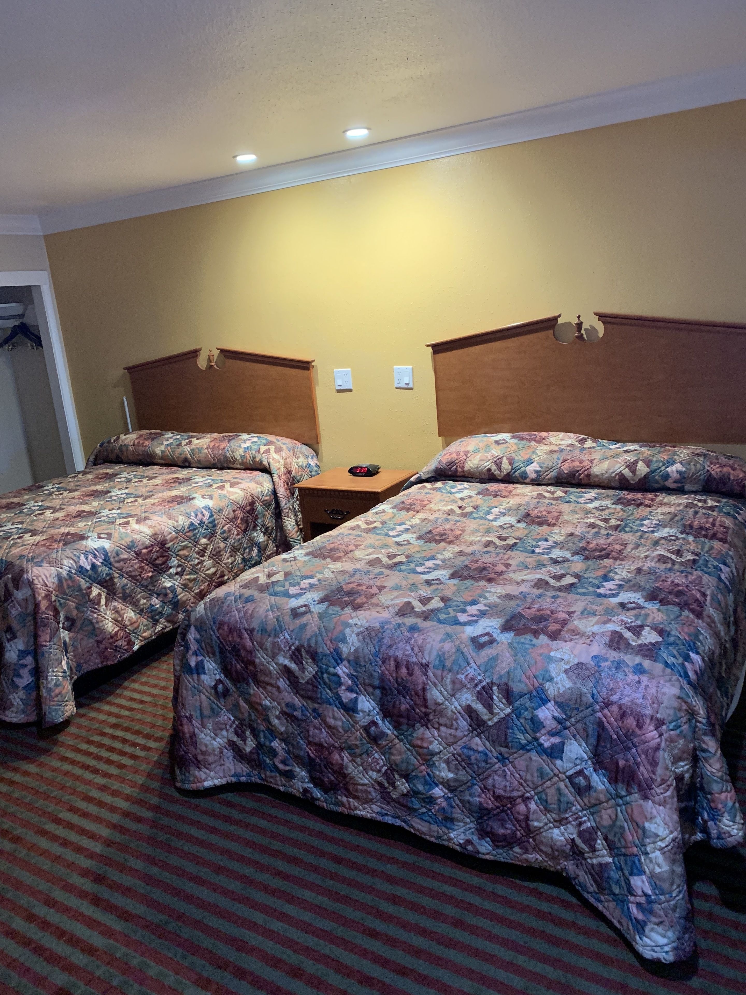 Habitación de hotel con dos camas, cada una con colcha de estampado floral, cabecero de madera y una mesita con un reloj entre ellas.