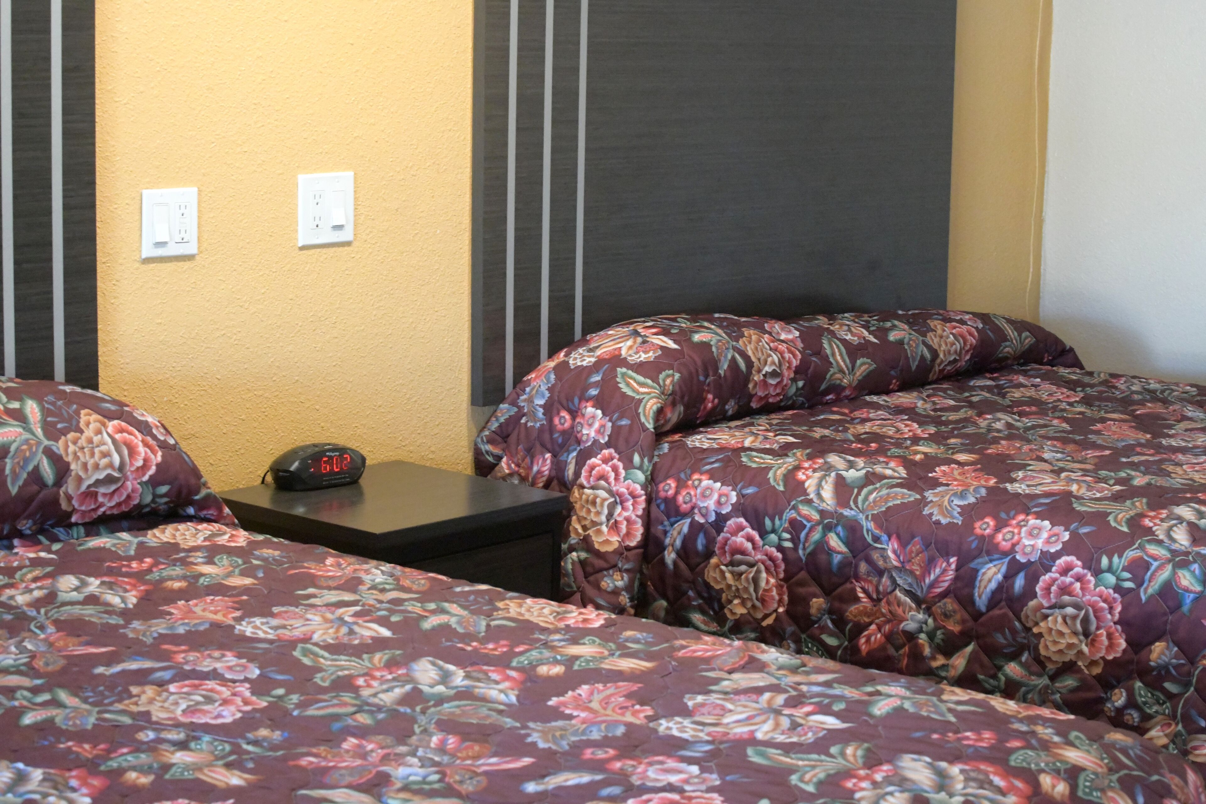 A hotel room with two beds, each with floral-patterned bedding, a small bedside table with a clock, and two electrical outlets on the wall.
