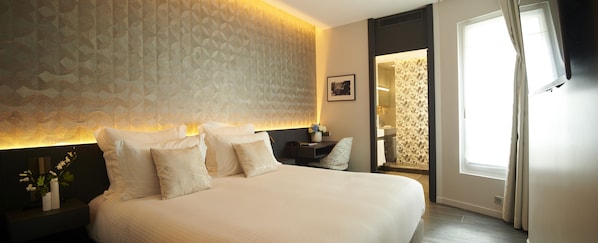 A modern hotel room with a large bed, a bedside table with a vase of flowers, a desk, and a view of the bathroom. - Roi de Sicile Rivoli (Paris)