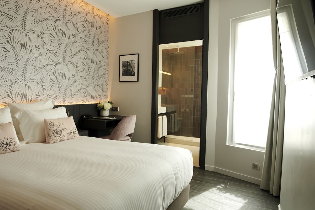 A bedroom with a large bed, a nightstand with a vase of flowers, a wall with a leaf pattern, and a bathroom with a shower and a towel rack.