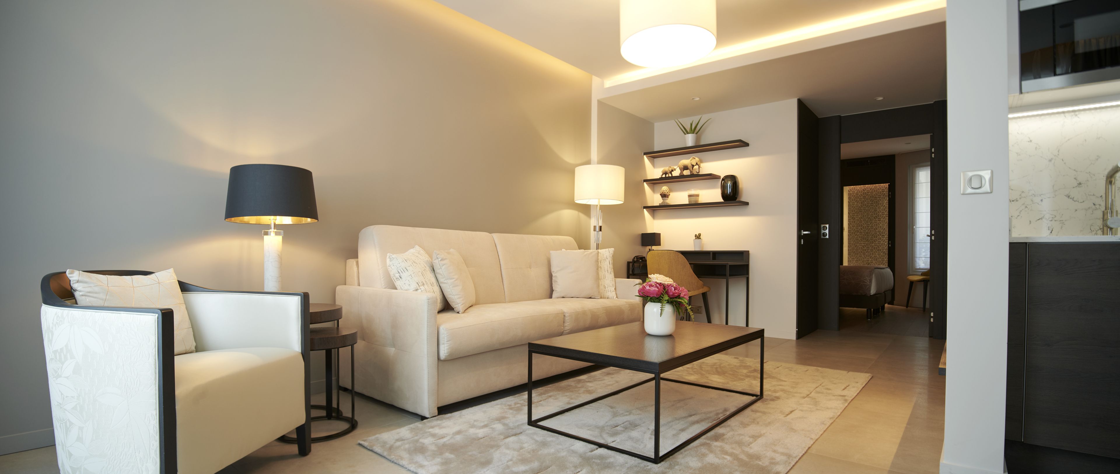 A modern living room with a sofa, armchair, coffee table, and a lamp. There is a kitchen area visible in the background.