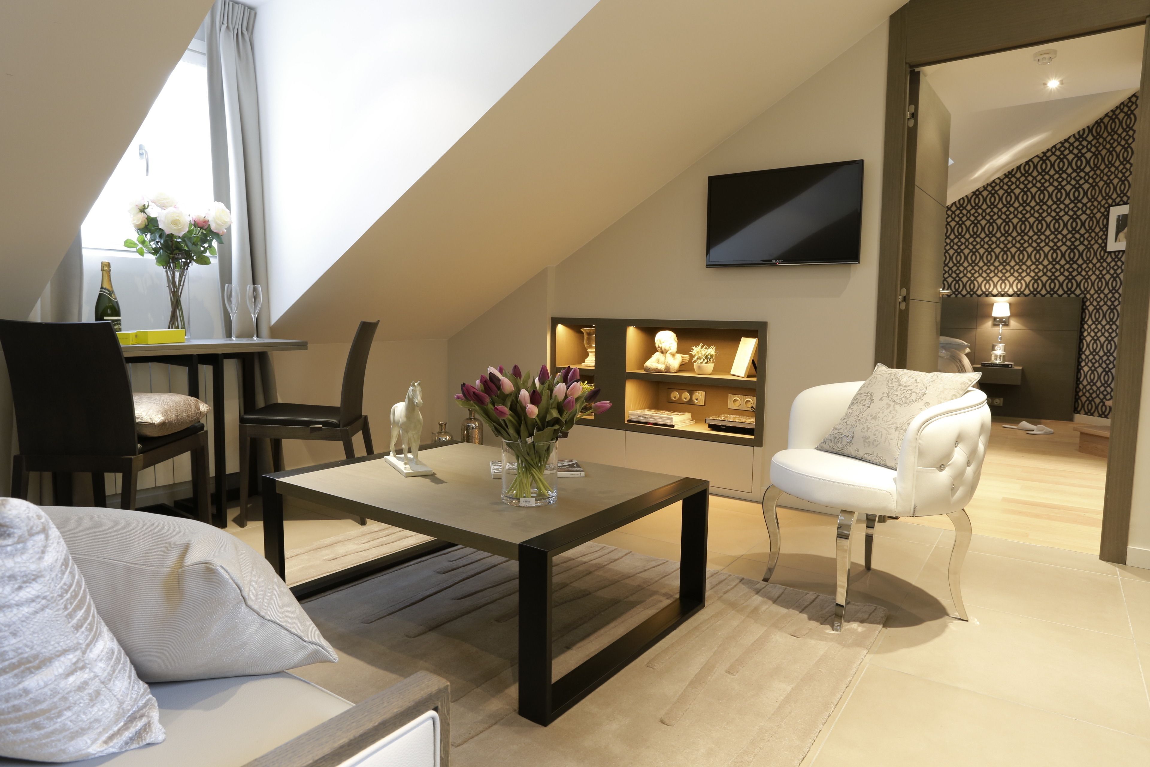 A modern living room with a dining area, a flat-screen TV mounted on the wall, and a small kitchen area with a TV and decorative items.