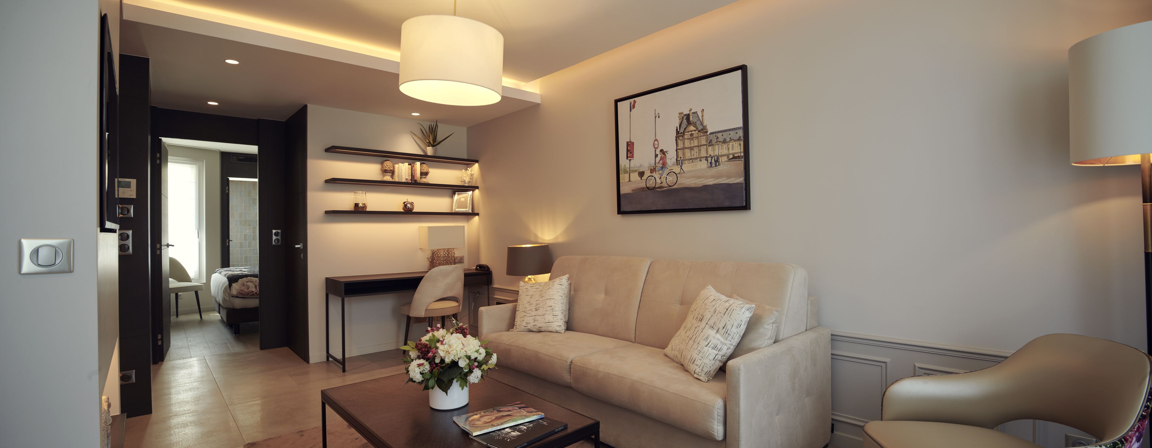 Un salon moderne avec un canapé, une table basse et un tableau représentant un bâtiment. On y trouve également un bureau avec une chaise et des étagères au-dessus.