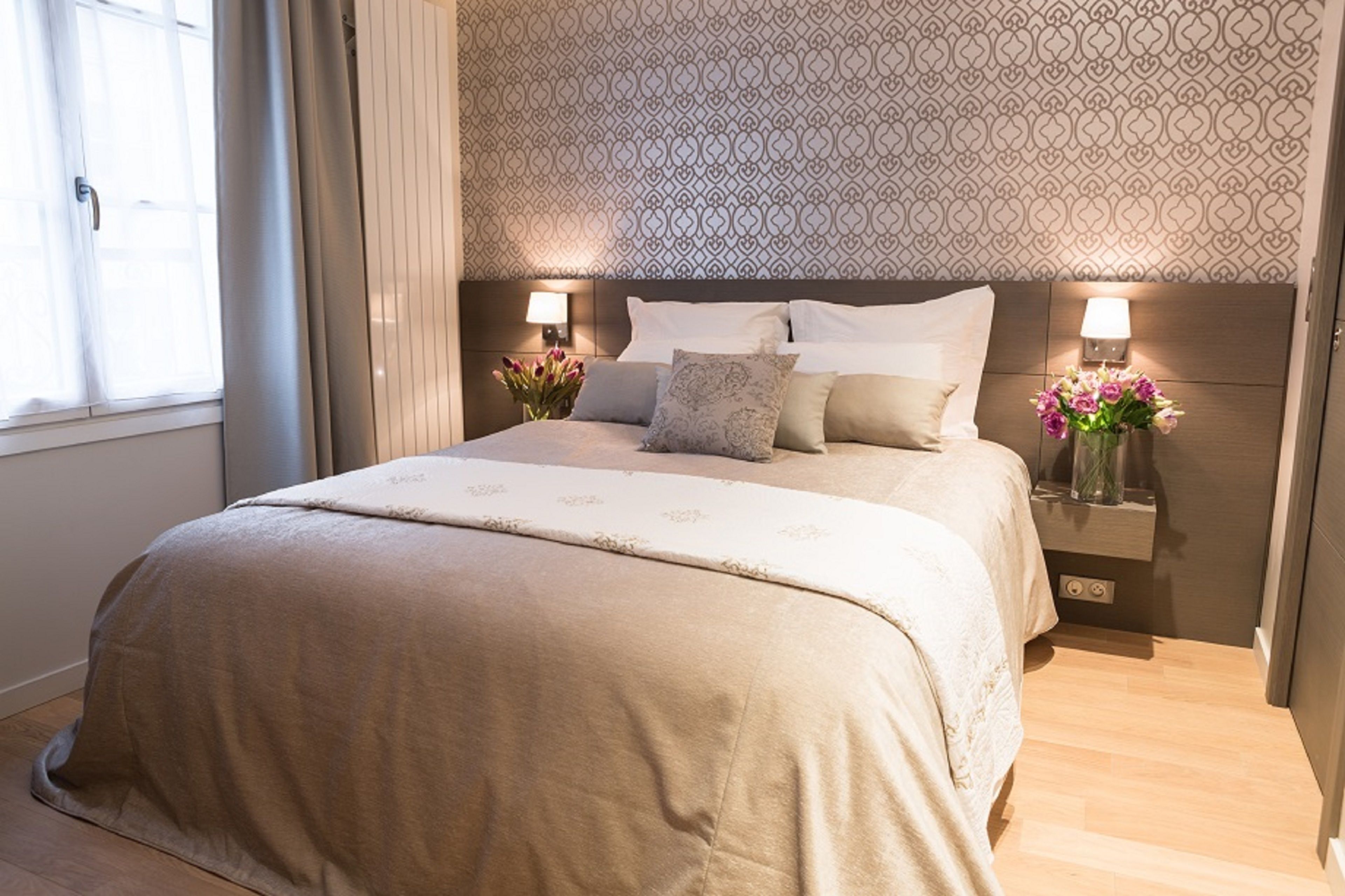 Un lit bien fait, avec une couvre-lit beige et des oreillers, deux lampes de chevet et un vase rempli de fleurs sur la table de chevet.
