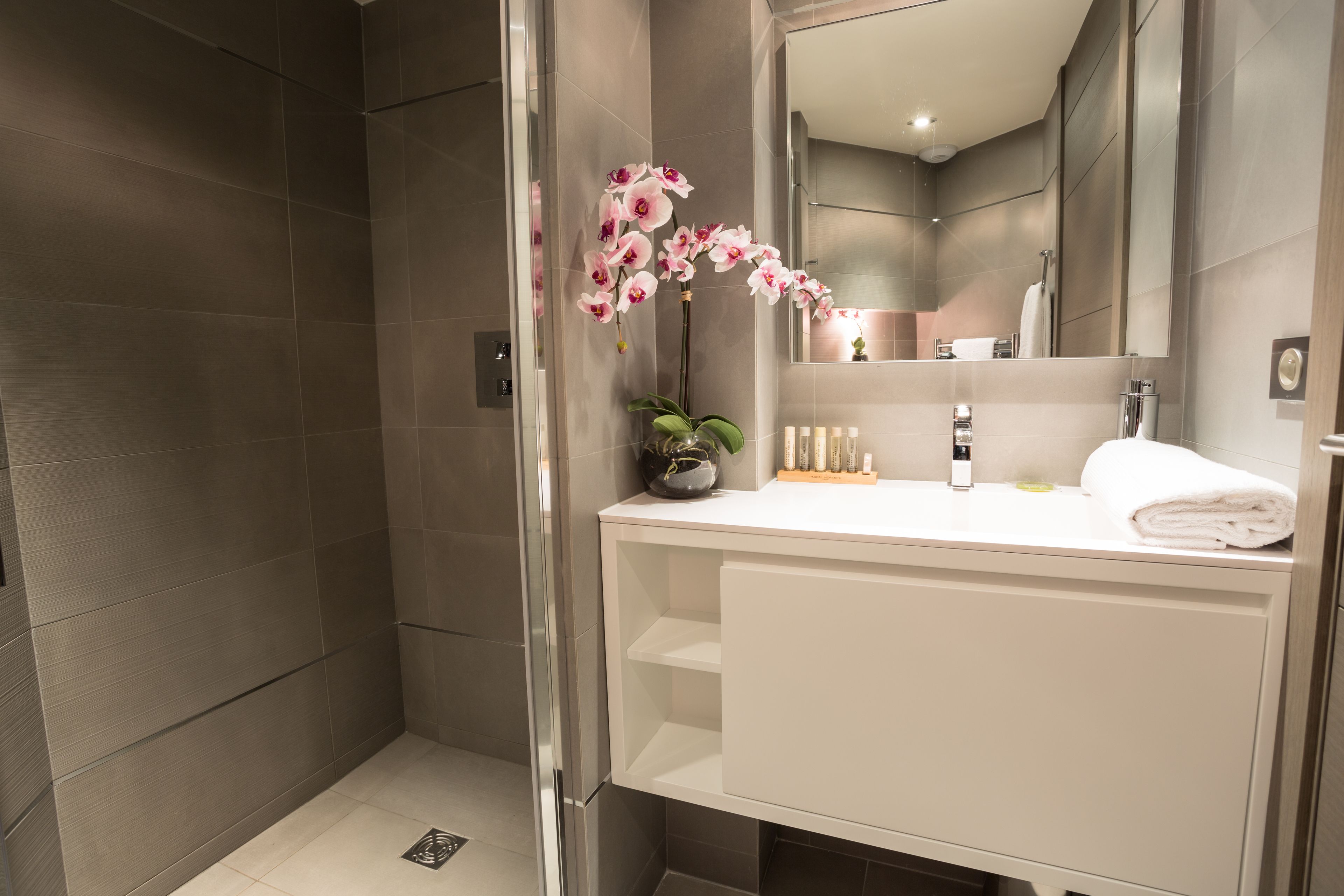 Un baño moderno con cabina de ducha de vidrio, tocador blanco con espejo y una orquídea en maceta.